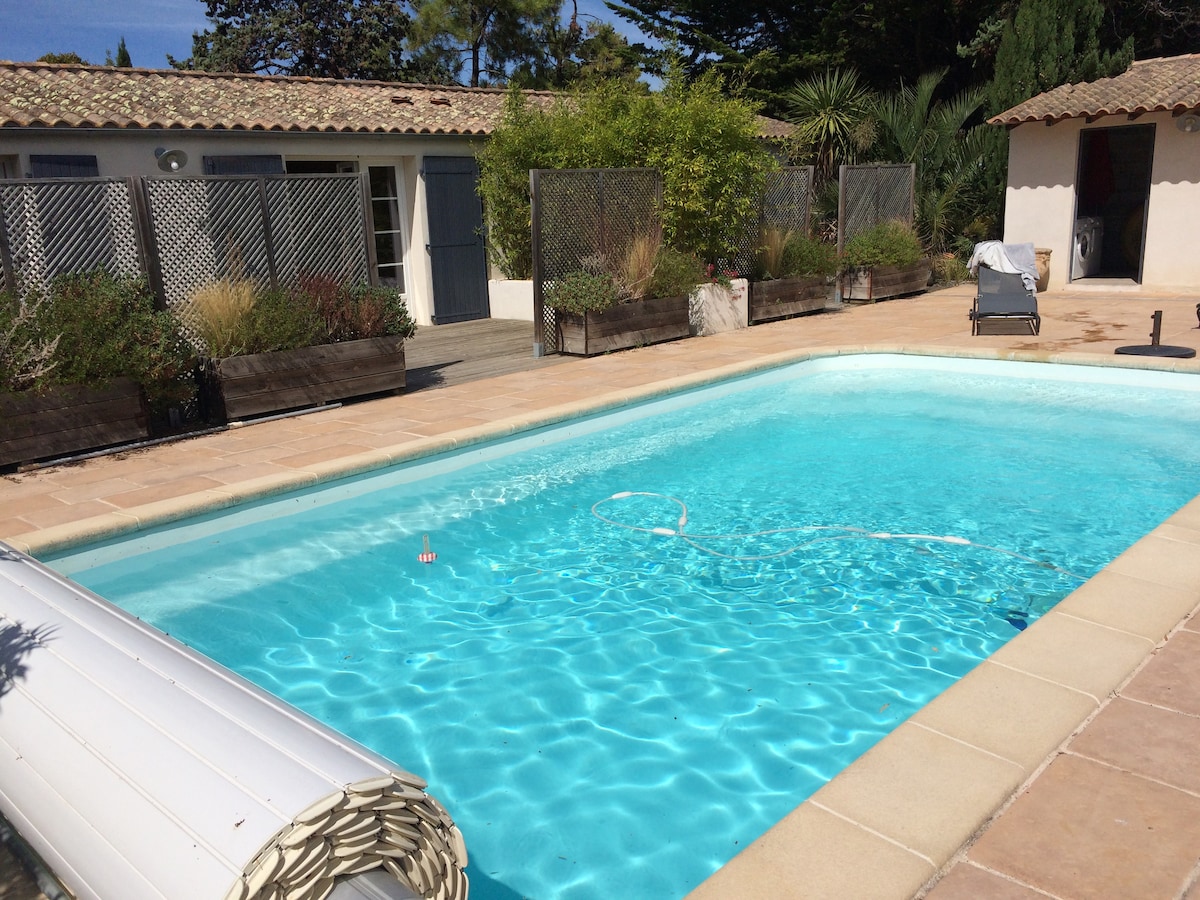 A clear blue swimming pool is surrounded by landscaped gardens and wooden planters. The area features white lounging chairs and a tranquil setting, with nearby buildings visible in the background, providing an inviting atmosphere for relaxation.