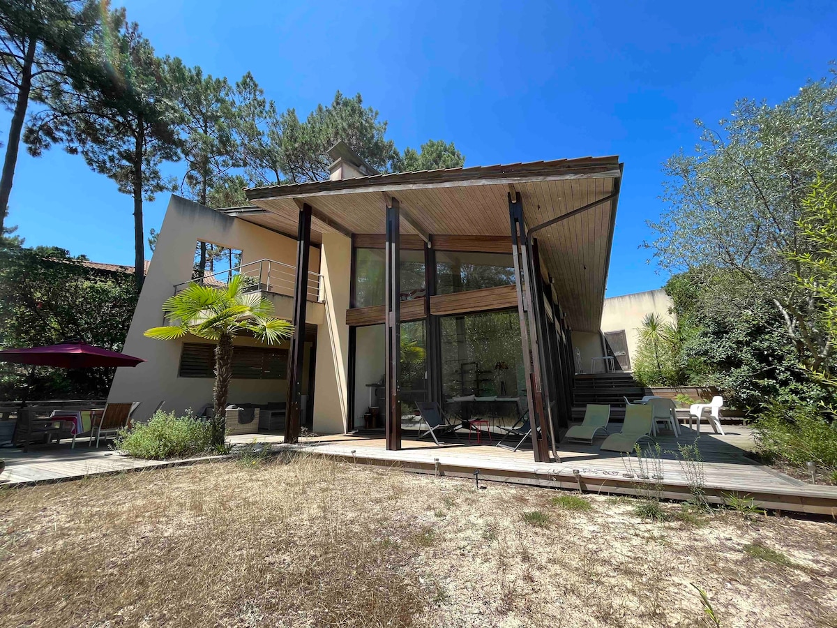 A modern architectural home features large glass windows reflecting natural light, surrounded by greenery. The exterior is complemented by wooden beams, while a patio area with outdoor furniture is visible in front. A hint of a landscaped garden can be seen in the foreground.