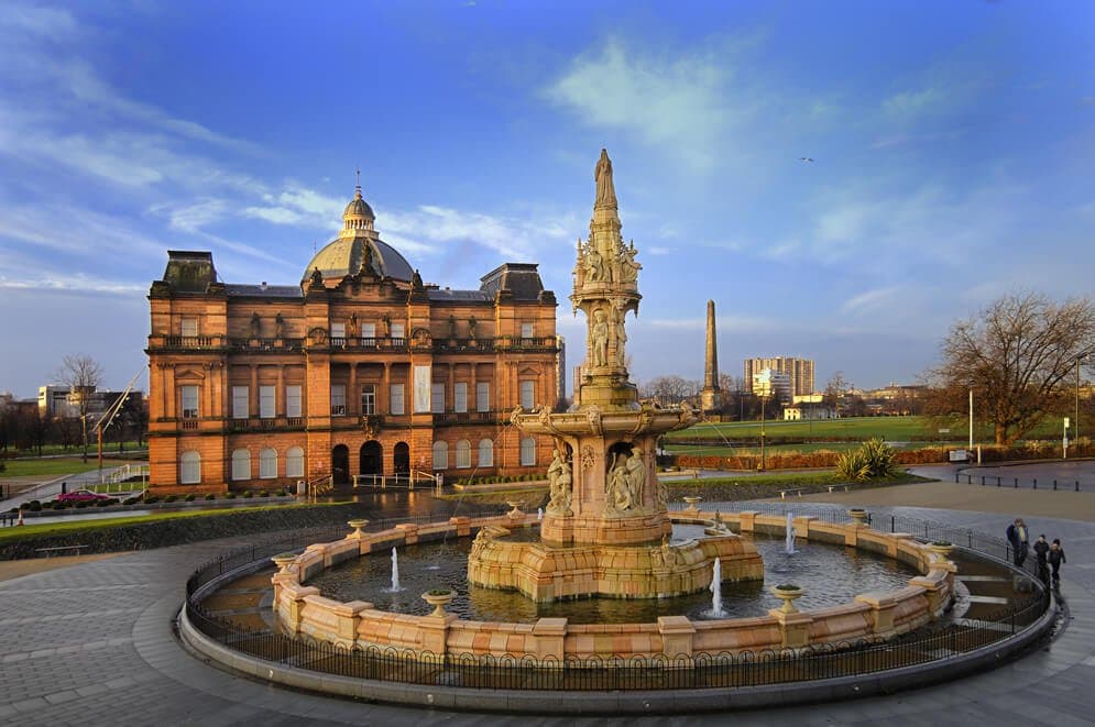 The image captures a grand historical building with ornate architecture, set against a vibrant sky. In the foreground, a large fountain features intricate sculptures and water spouting gracefully. Nearby, landscaped gardens and pathways create an inviting atmosphere for leisurely walks.