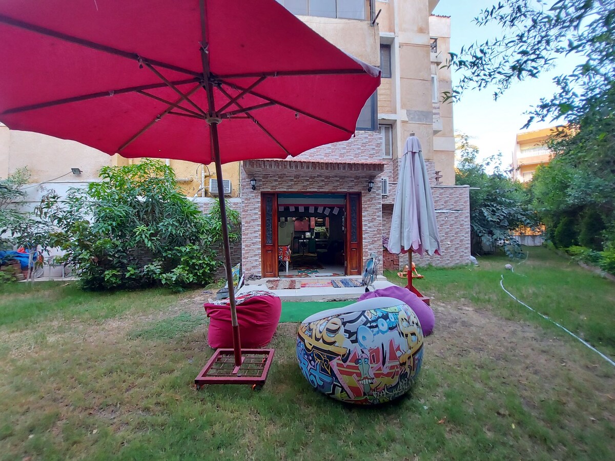 A private garden is showcased, featuring a large red umbrella and colorful bean bags. The lush grassy area is surrounded by greenery, with a brick wall and an open entrance to the interior visible in the background. A white patio umbrella adds to the outdoor seating arrangement.