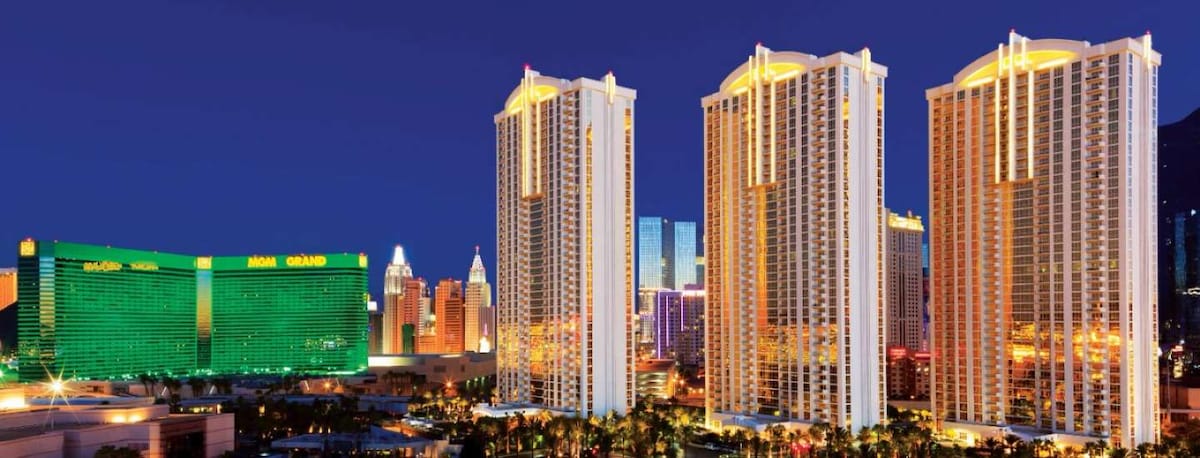 The image captures the elegant architecture of two high-rise towers glimmering at night. Surrounded by vibrant city lights, the buildings feature illuminated facades and a well-maintained landscape, creating a striking view of the Las Vegas skyline.