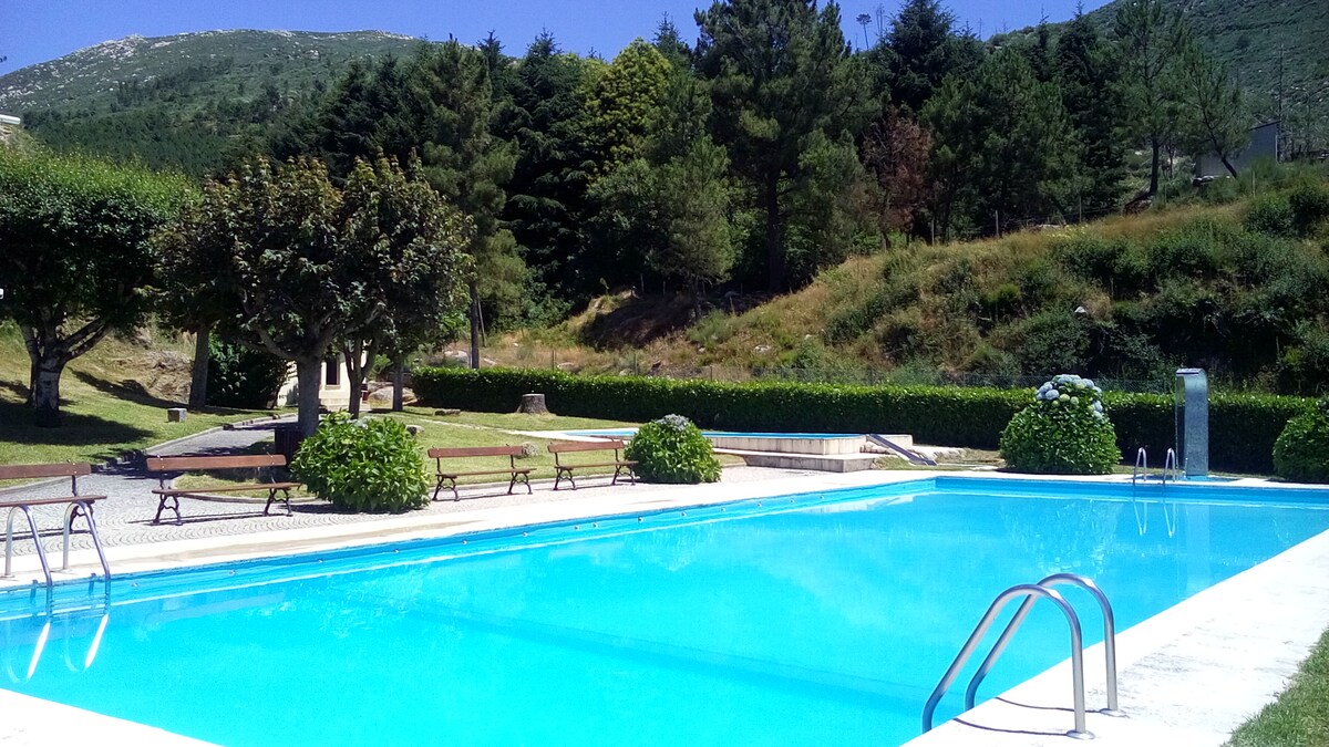 A clear blue pool is set within a landscaped area, surrounded by greenery and trees. Wooden benches are positioned around the pool, offering spaces to relax. The background features gentle hills, enhancing the tranquil atmosphere of the outdoor space.