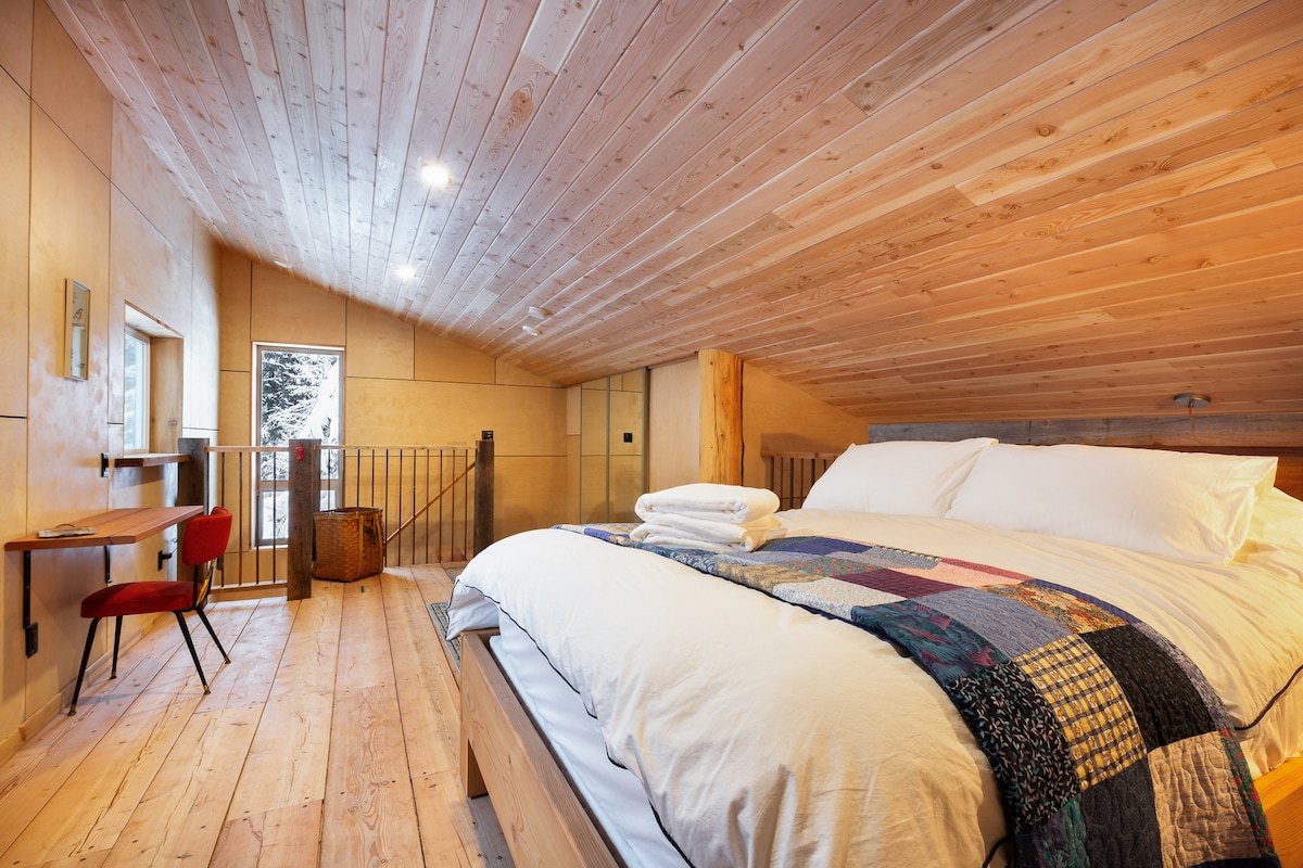 An airy loft space features a spacious bed adorned with a patchwork blanket. Natural wood beams and flooring create a warm ambiance. A small desk with a red chair sits adjacent to a window, allowing natural light to fill the room. Complementing the design is a nearby storage basket.