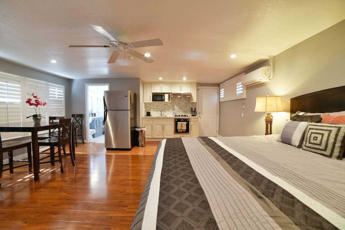 A remodeled studio space features a large bed with a geometric patterned blanket. A kitchenette is visible in the background, equipped with modern appliances. A dining table is set for two, and the space is well-lit by overhead lights and natural light from nearby windows.