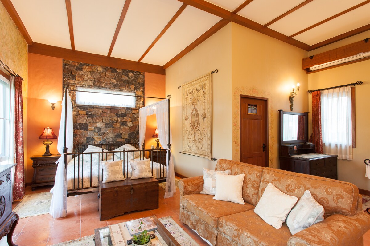 The spacious suite features a king-size bed with a four-poster design, complemented by decorative wall art and a stone accent wall. A comfortable seating area with a sofa is arranged in front of a coffee table, while a flat-screen TV and light filtering through sheer curtains enhance the inviting atmosphere.
