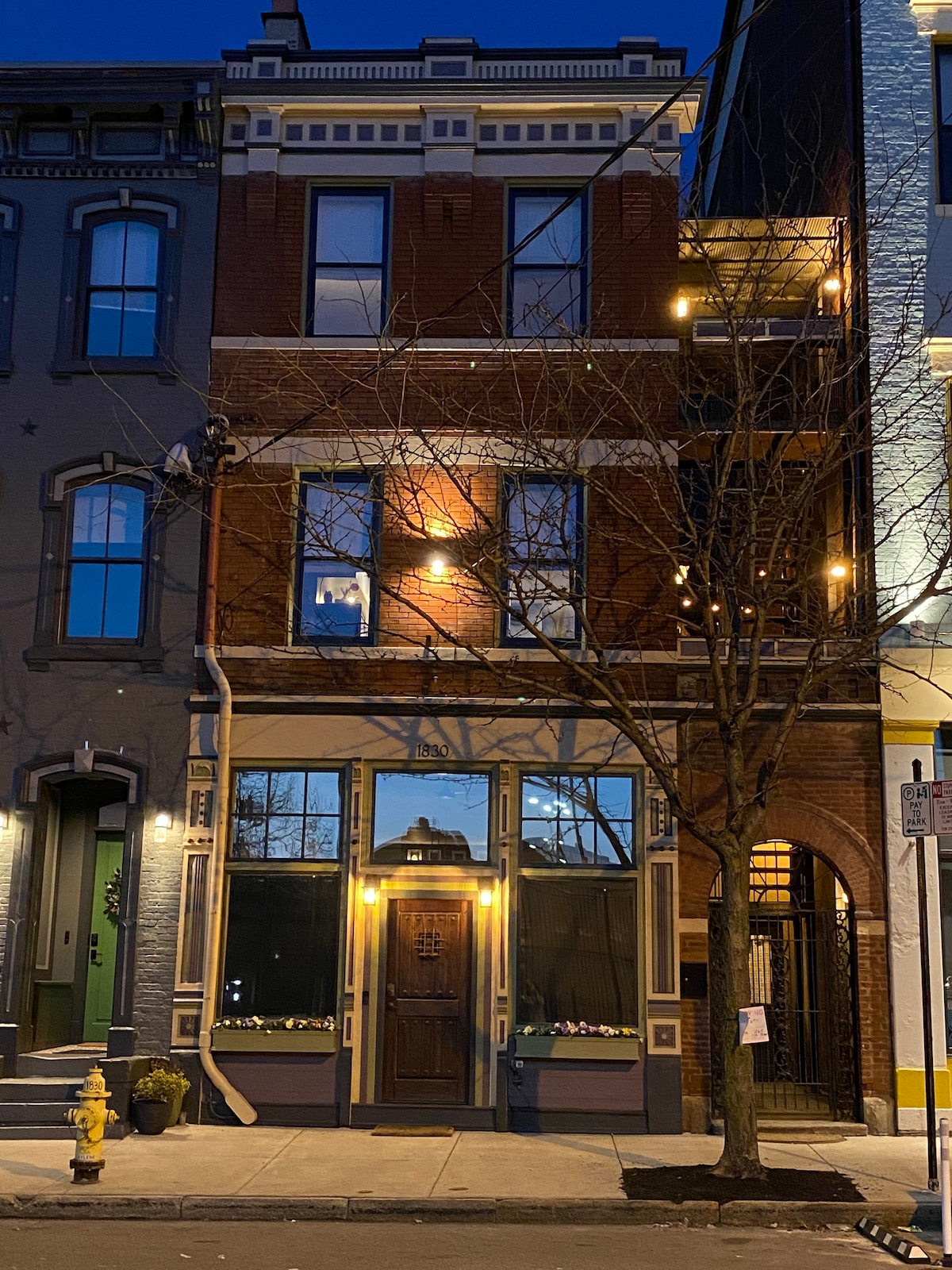 The exterior of a three-story historic building is showcased, featuring a brown door and multiple windows illuminated by warm lights. A tree with bare branches stands in front, and the surrounding architecture reflects the character of the neighborhood.