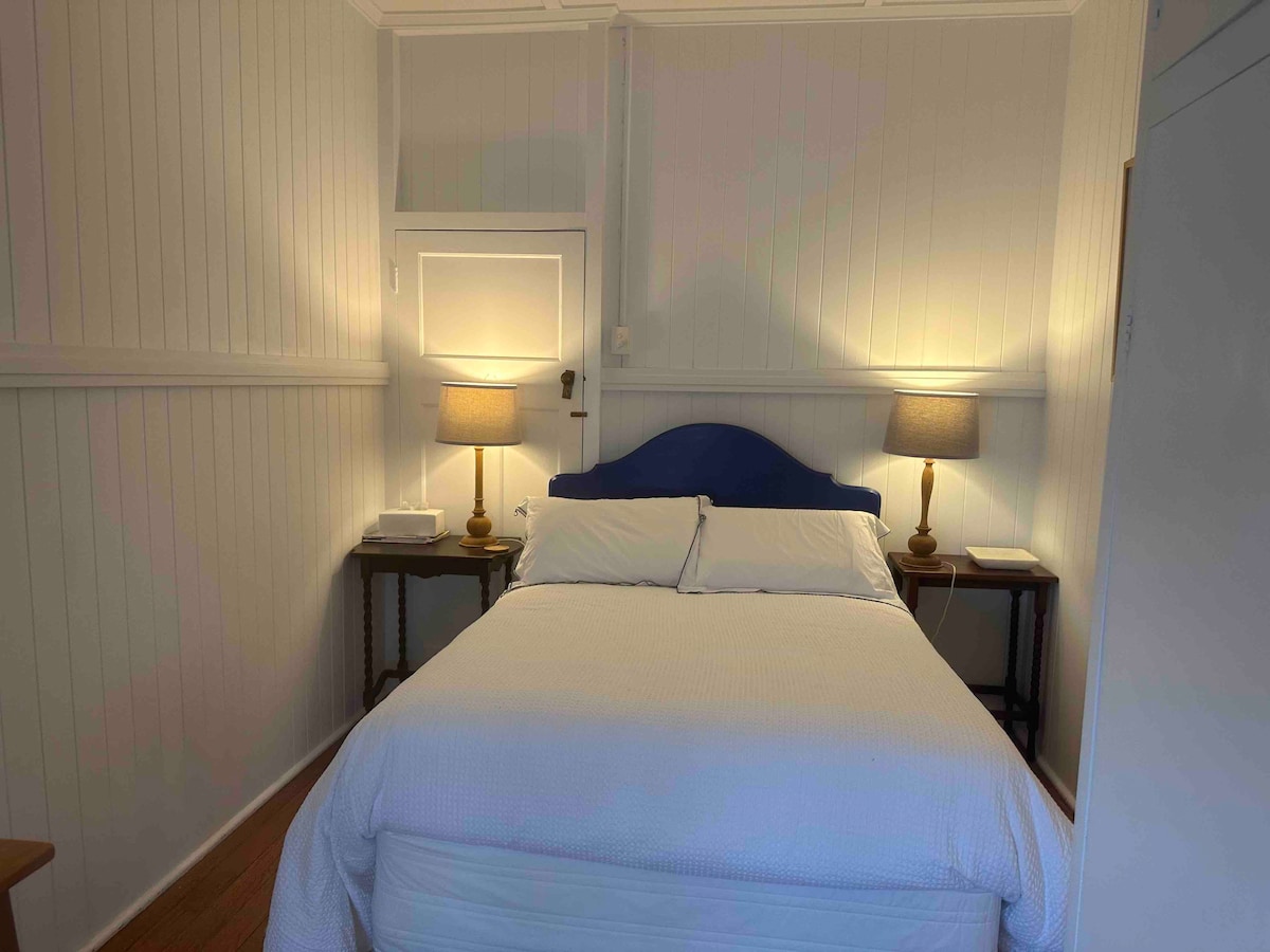 A cozy bedroom is furnished with a spacious bed, complemented by bedside tables with lamps on either side. Gentle illumination enhances the soft texture of the bedding and the light-toned wooden walls, creating a restful environment.