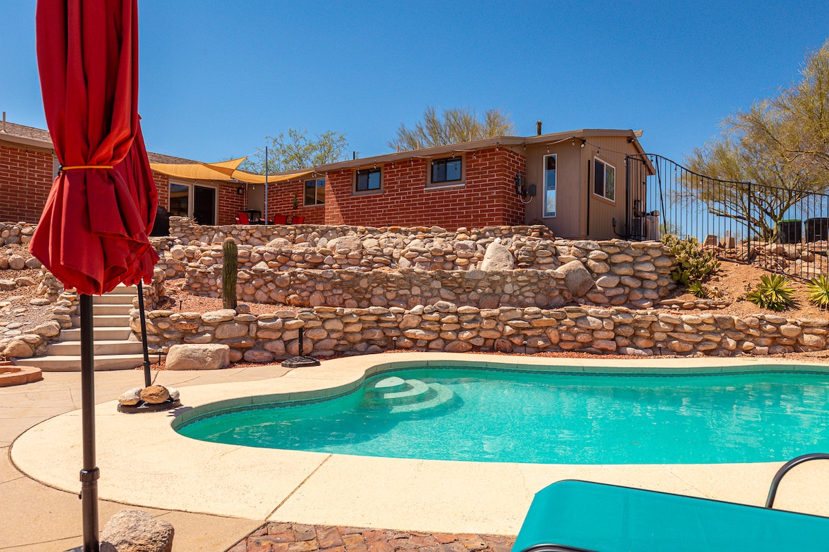A private pool is surrounded by a well-maintained patio, offering lounge chairs and a shaded umbrella. Stone landscaping creates visual interest, and desert vegetation enhances the natural setting. The studio and surrounding structures are visible in the background against a clear blue sky.