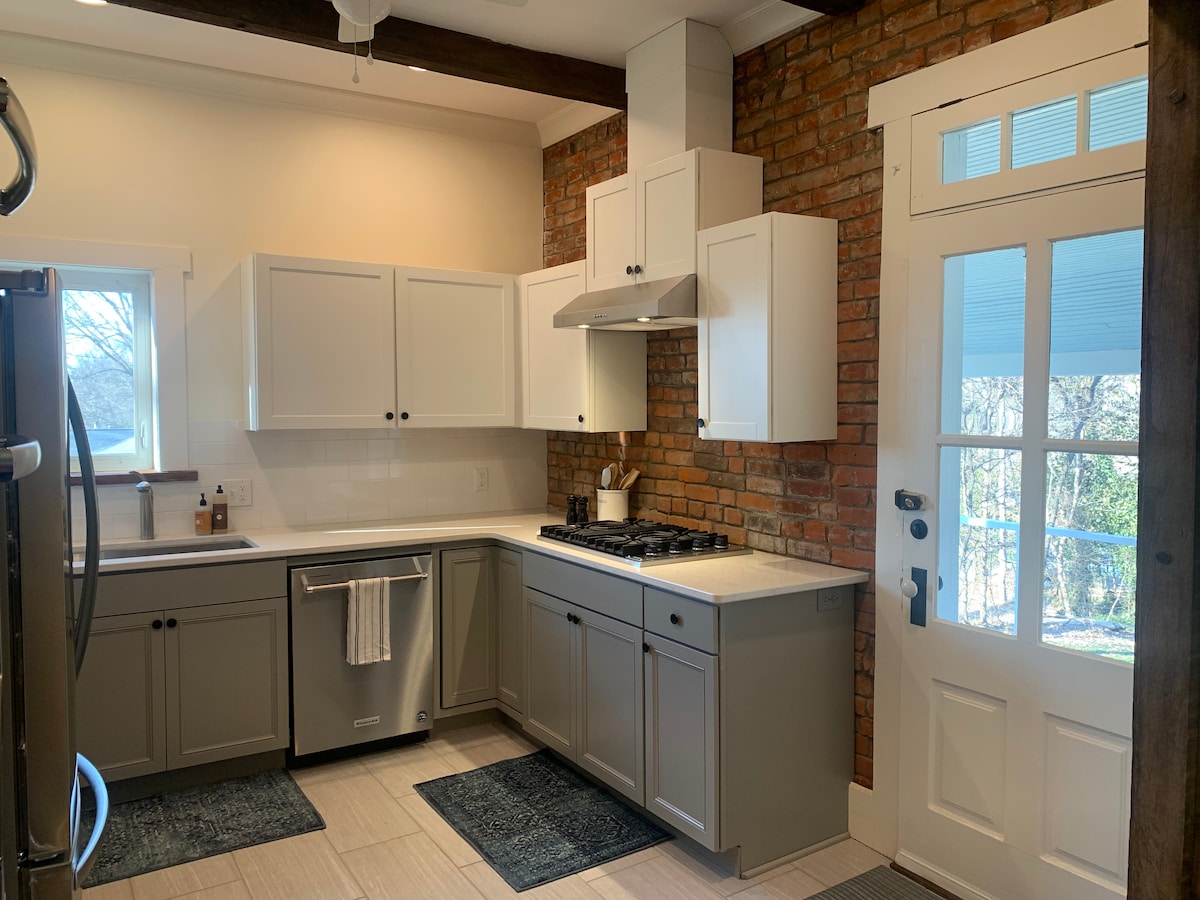 The kitchen features modern cabinetry with a blend of white and gray finishes, complemented by exposed brick walls. Essential appliances, including a stainless steel refrigerator and a gas range, are present. Natural light enters through a window, enhancing the functional layout.