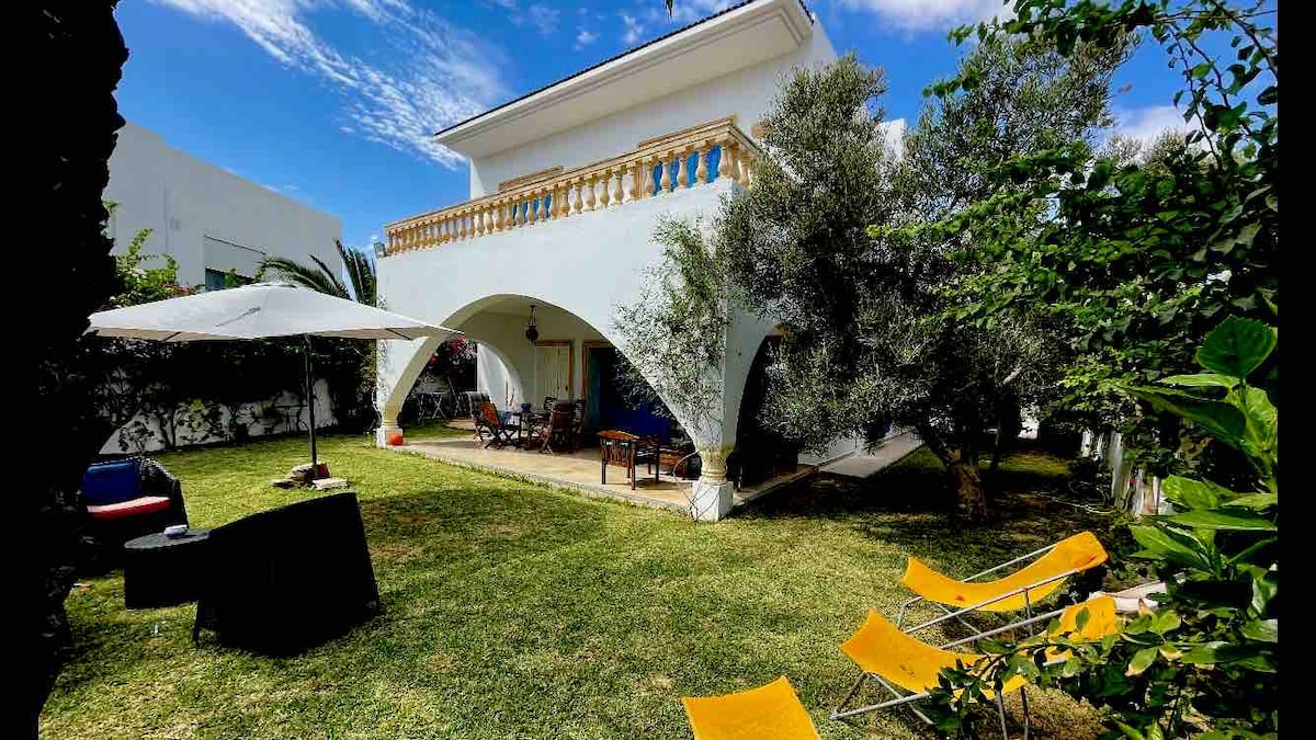 The exterior of the villa features a spacious garden with manicured grass, surrounded by lush greenery. An outdoor seating area provides shade under a large umbrella, while several lounge chairs are positioned for relaxation. The balcony above overlooks the inviting space.