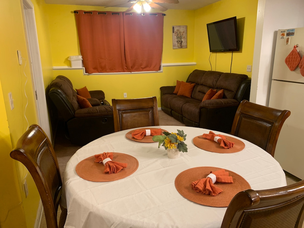 A bright dining area features a round table set with decorative placemats and neatly folded napkins. Comfortable seating includes a dark sectional sofa and an armchair, positioned near a wall-mounted TV. The walls are painted a vibrant yellow, enhanced by rich orange curtains.