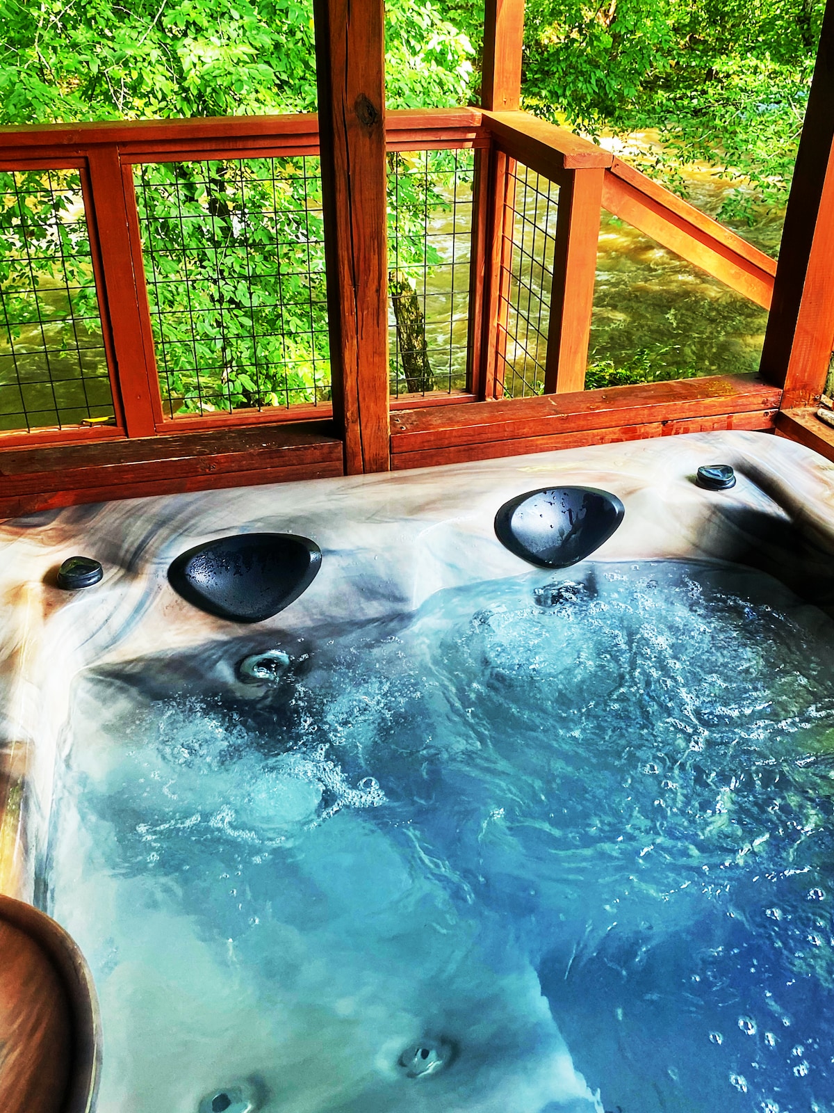 A private hot tub is situated on a wooden porch, surrounded by a railing that reveals lush greenery and the flowing water of a nearby creek. Bubbles are visible within the water, indicating a relaxing atmosphere.