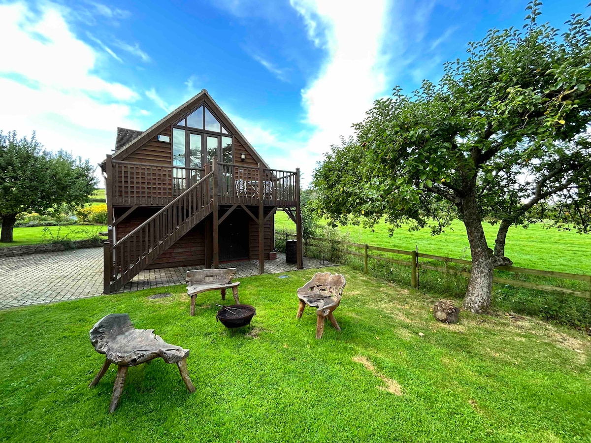 A wooden structure with a second-story balcony is surrounded by lush green grass. Unique log furniture, including a fire pit, is arranged in a natural setting. A tree offers shade, and open fields extend beyond the property, under a clear blue sky.