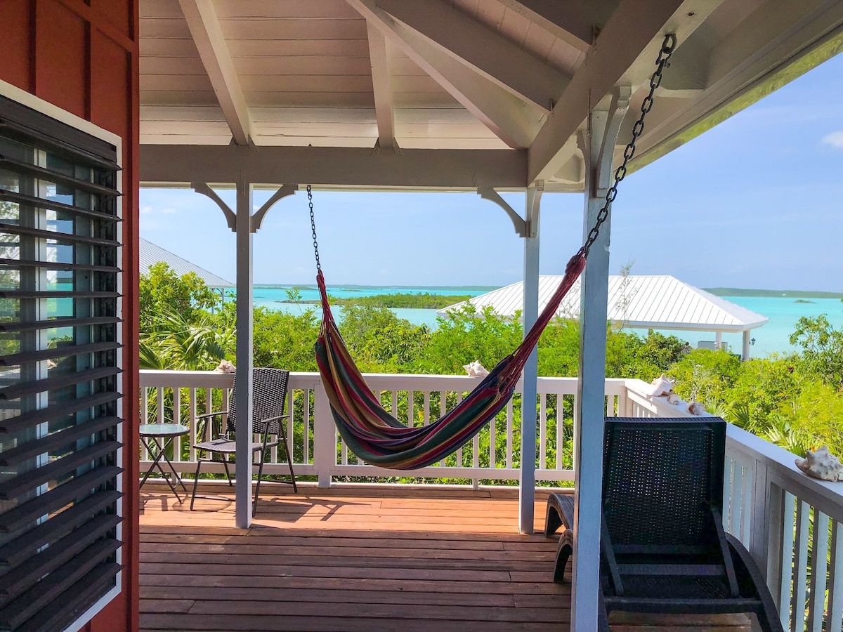 A private balcony is featured, adorned with a colorful hammock suspended from a wooden beam. Chairs are positioned for relaxation, with views of the lush greenery and distant turquoise waters of Chalk Sound visible beyond. The area is sheltered by a pitched roof.