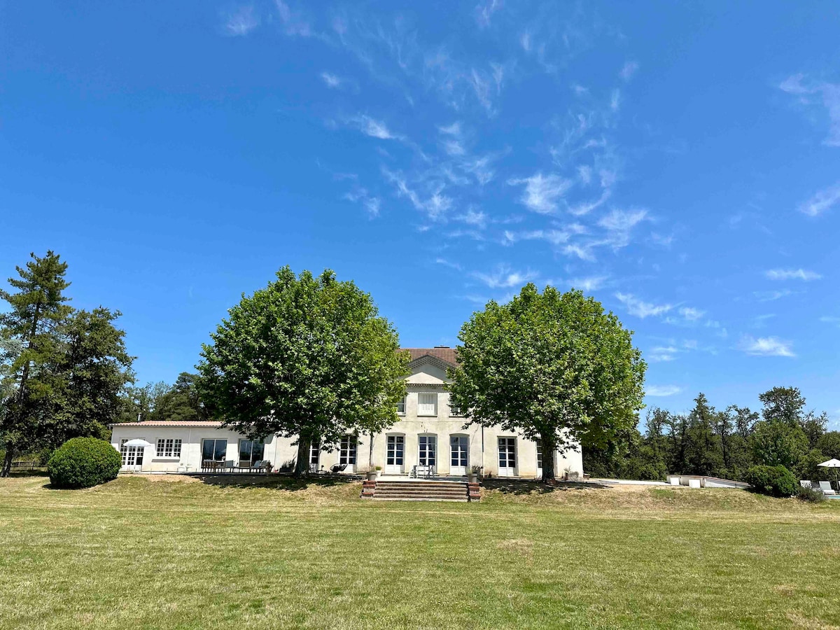 The exterior of a spacious family home is showcased, framed by mature trees. The house is presented with a large lawn area in front, featuring bright blue skies and wispy clouds overhead. The entrance is marked by a set of steps leading to the main door.