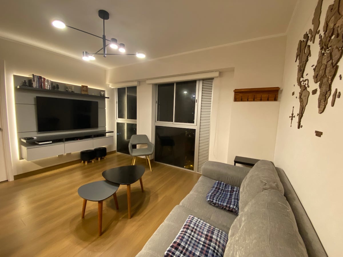 A modern living area features a grey sofa with throw pillows, paired with a set of wooden coffee tables. A large flat-screen TV is mounted on a sleek cabinet, and two windows allow for natural light to fill the space. Decorative wall art adds character.