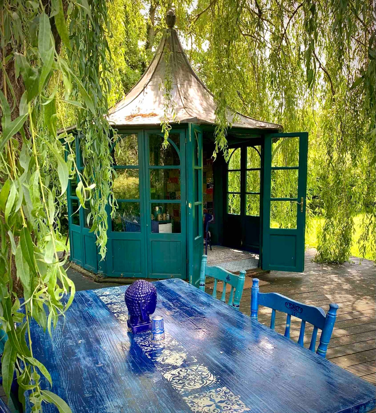 A charming gazebo is framed by the soft draping of willow branches, with turquoise walls and glass doors inviting exploration. A rustic blue table sets the scene outside, complemented by decorative accents, creating an inviting space for gatherings or quiet reflection.