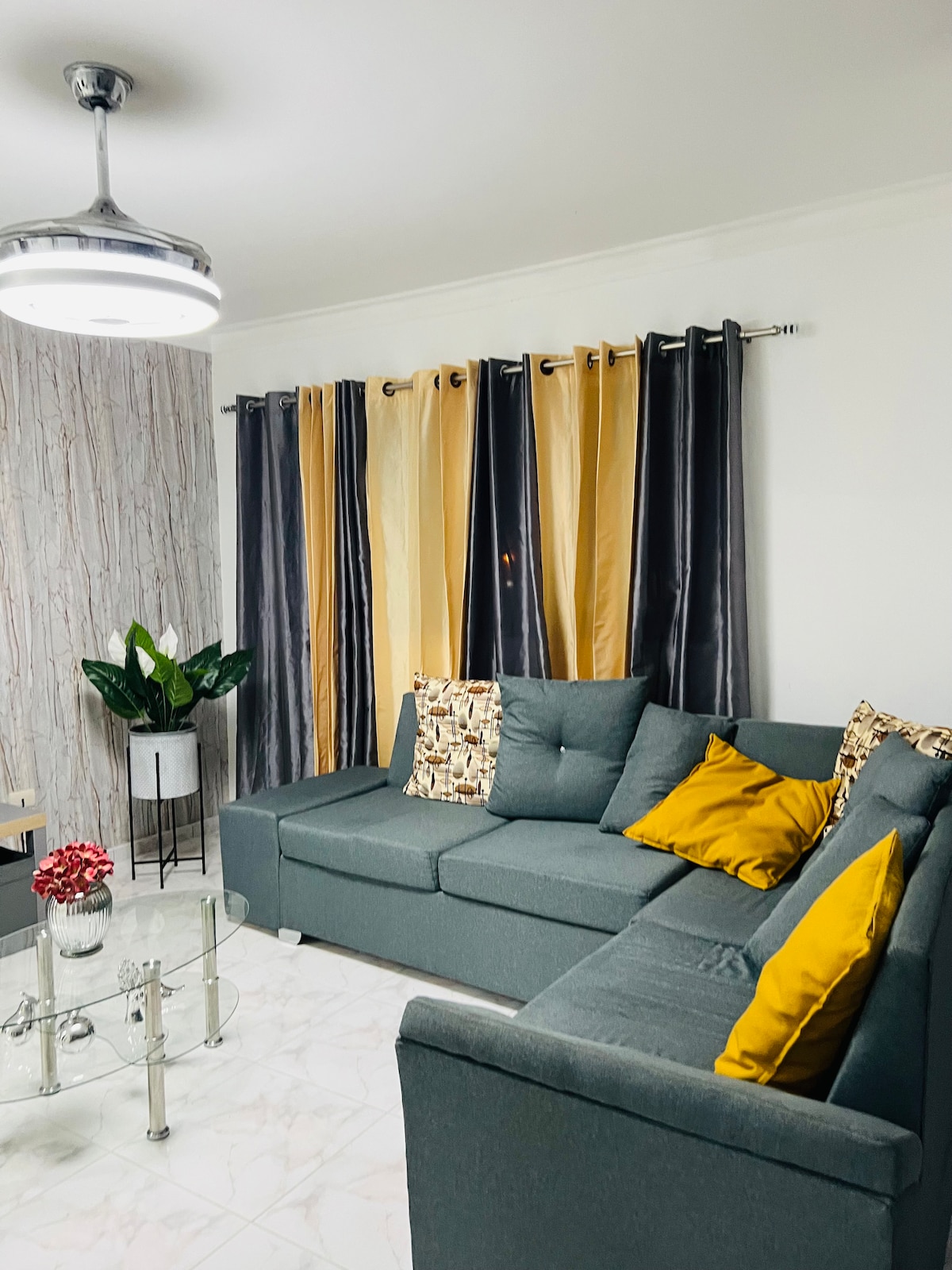 A comfortable living area features a gray sectional sofa with decorative cushions in yellow and patterned fabric. A glass coffee table is positioned in front of the sofa, complemented by a potted plant. Floor-to-ceiling curtains in gray and yellow frame a window, adding warmth to the space.