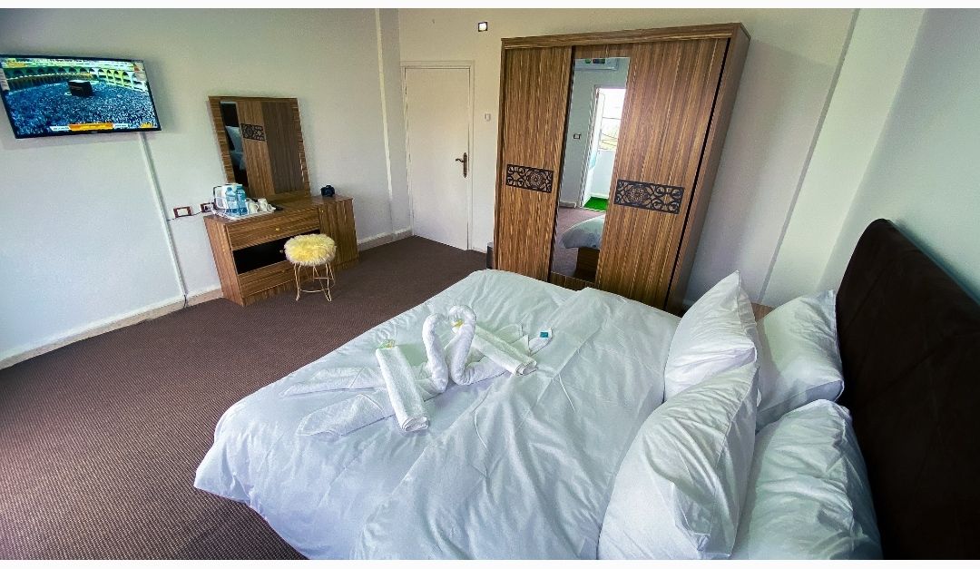 A double room features a spacious bed with neatly arranged linens. A wooden wardrobe and a vanity mirror are visible, as well as a modern television mounted on the wall. Soft carpeting enhances the comfort of the space.