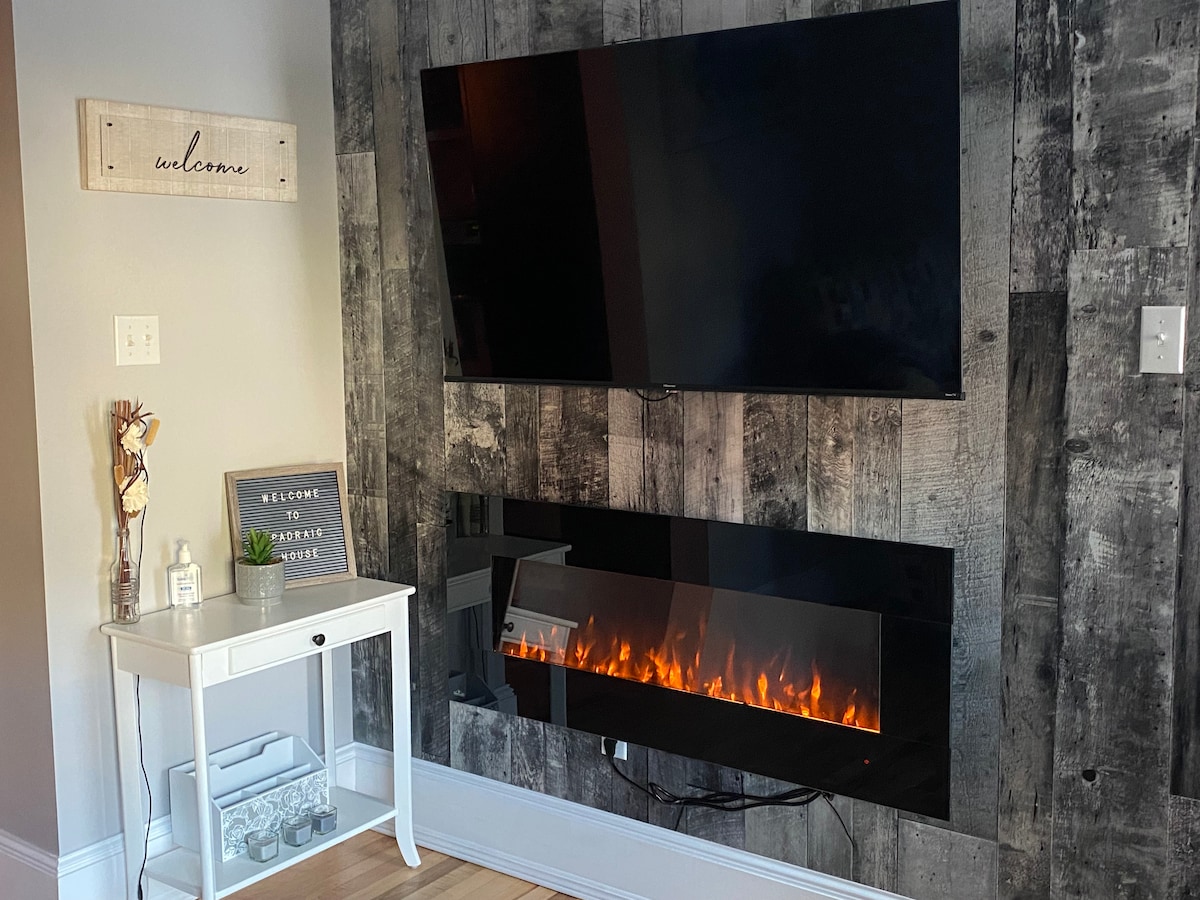 A modern living area features a mounted flat-screen TV above a sleek electric fireplace, providing a contemporary focal point. A white accent table is positioned beside the fireplace, adorned with decorative items, while a welcome sign adds a friendly touch to the rustic wooden wall.