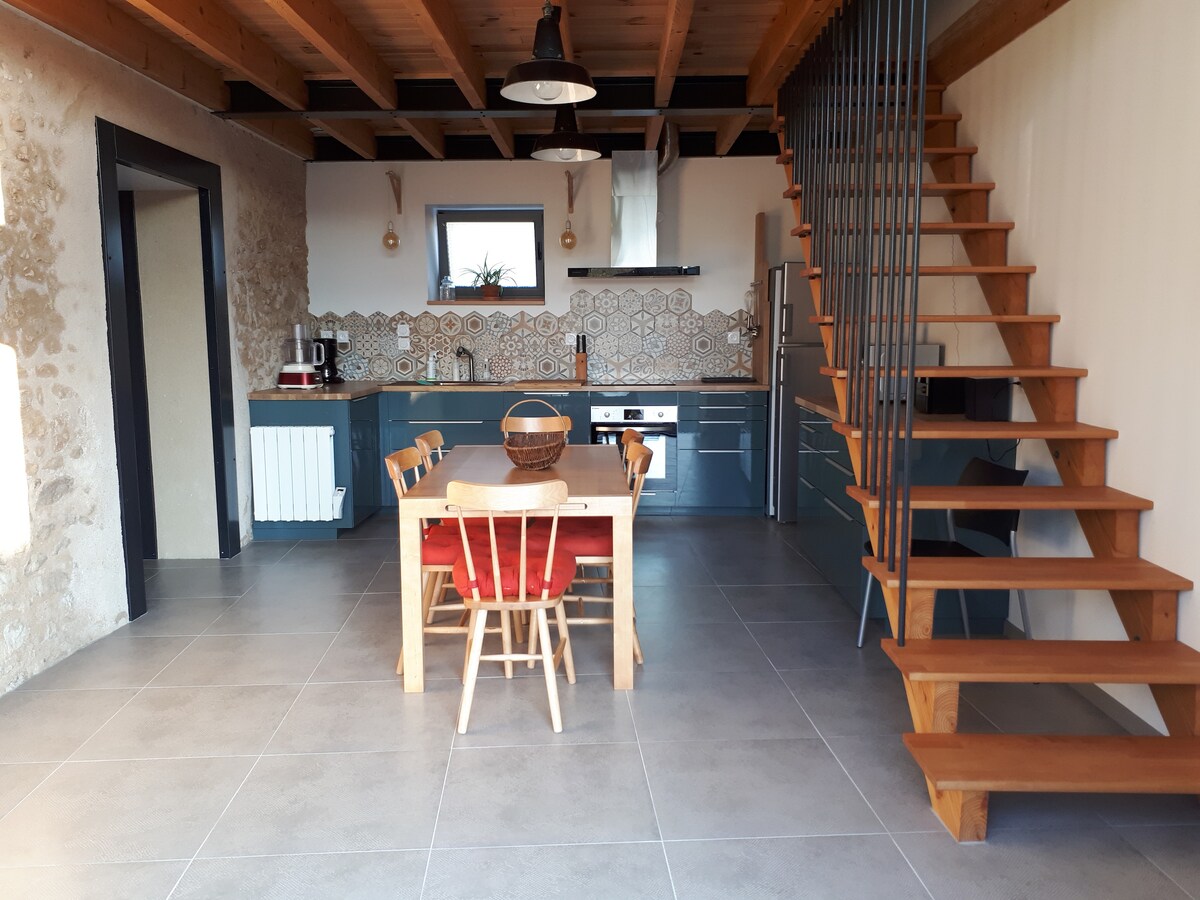 A spacious kitchen and dining area features a wooden table surrounded by four chairs. Modern appliances, including a refrigerator and oven, are visible in the kitchen. A staircase made of wood leads to the upper level, highlighted by stylish tiles along the wall.