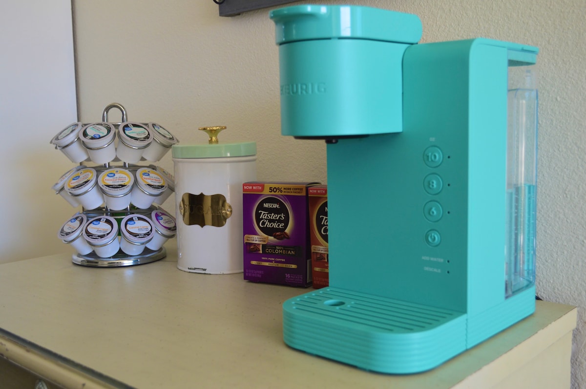 A turquoise Keurig coffee maker is positioned on a light-colored surface, beside a white canister with a gold lid. Multiple coffee pods are organized in a circular stand, featuring various brands, while boxes of instant coffee are displayed nearby.