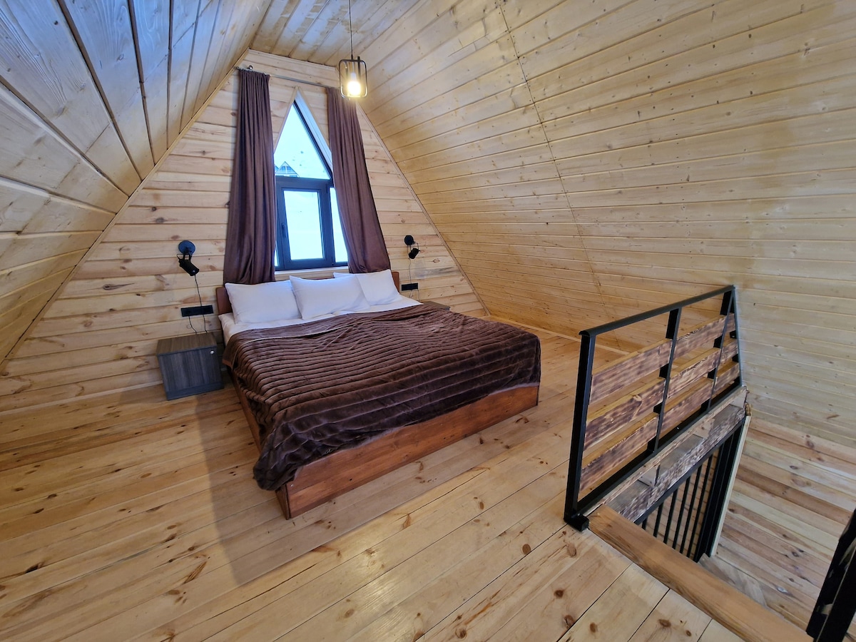 An inviting space features a spacious bed draped in a brown blanket. The wooden walls and ceiling create a cozy atmosphere, complemented by a large window allowing natural light to fill the room. Light fixtures are mounted on the walls, enhancing the design.