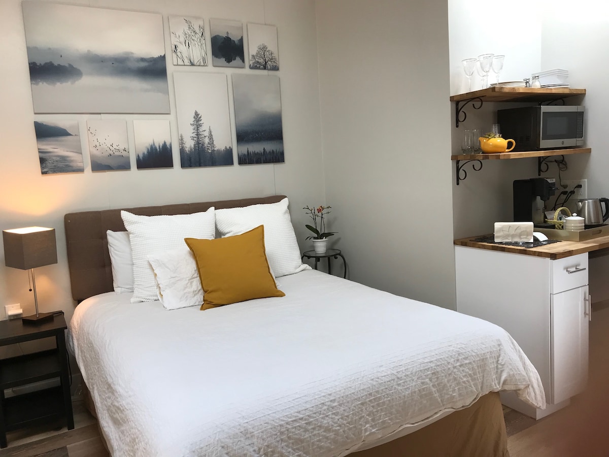 A comfortable sleeping area is presented with a spacious bed featuring white linens and a yellow accent pillow. Wall art depicting serene landscapes is displayed above the bed. A small kitchenette with a microwave and coffee bar is located in the corner, adding functionality to the space.