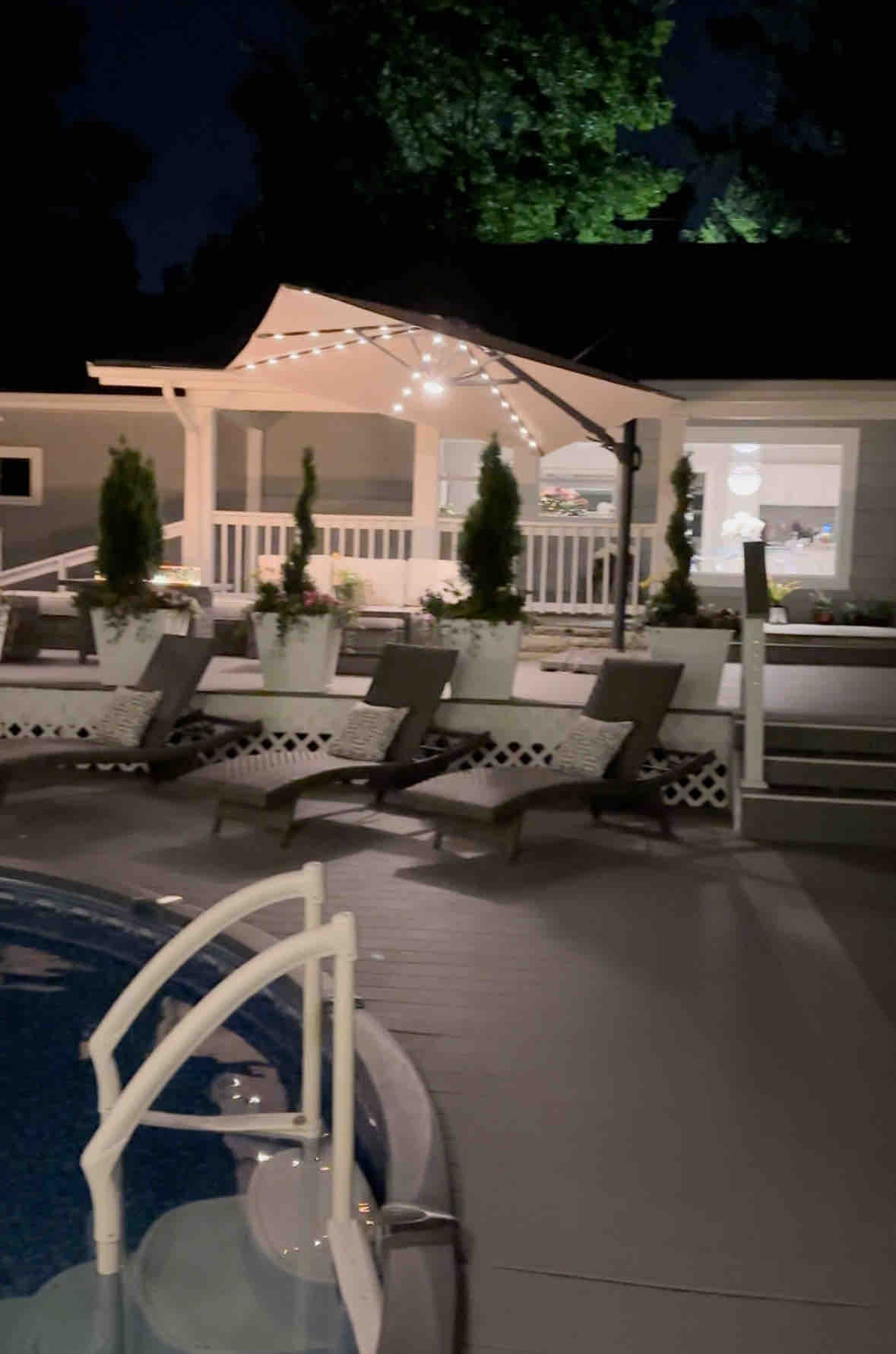 A cozy outdoor space is illuminated at night, featuring a covered seating area with string lights. Several lounge chairs are arranged beside a circular pool, creating an inviting atmosphere for relaxation.