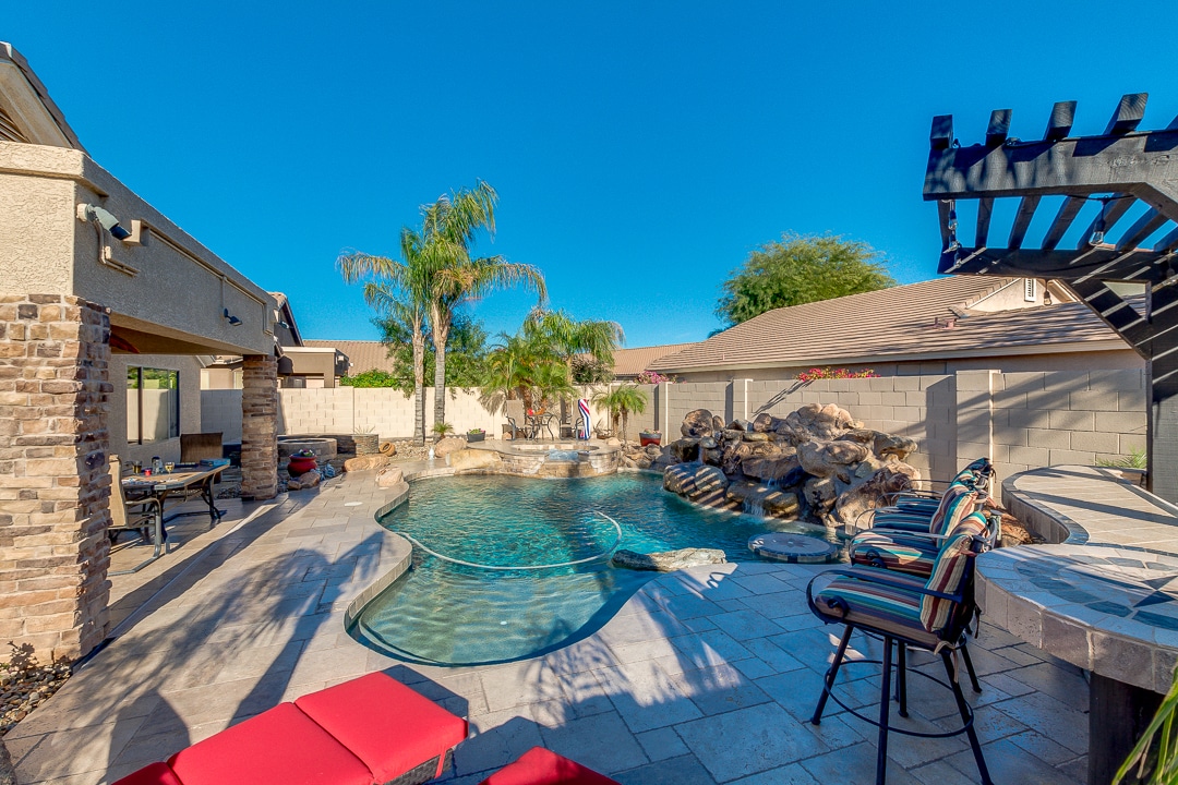A spacious outdoor area is presented with a swimming pool featuring a rock waterfall. Lounge chairs are positioned near the pool, and a shaded barbecue area is visible. Palm trees provide a tropical feel, enhancing the inviting atmosphere of the backyard.