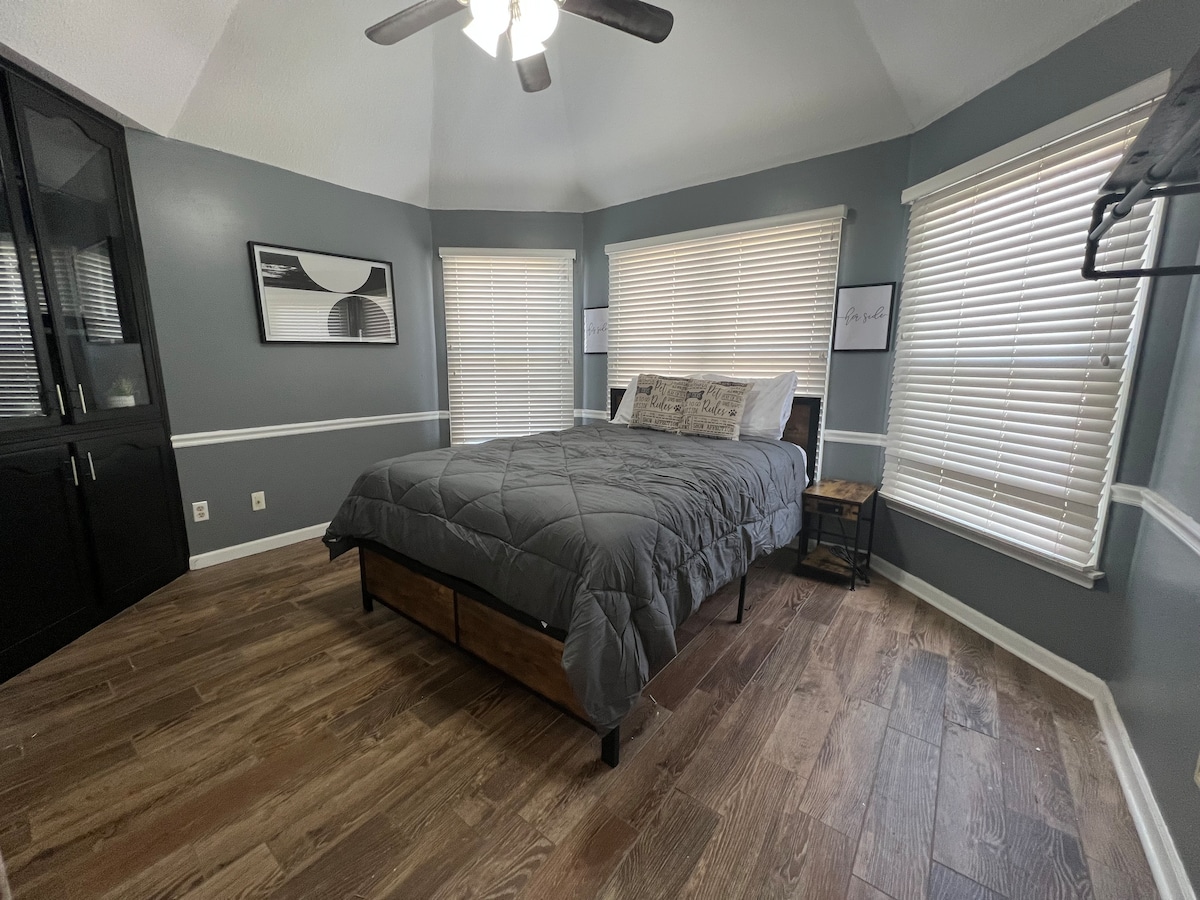 A calming bedroom features a spacious bed topped with a gray quilt. Natural light filters through two window shades, creating a bright atmosphere. A minimalist wall art piece is displayed above the bed, and dark wood flooring complements the soft color palette.