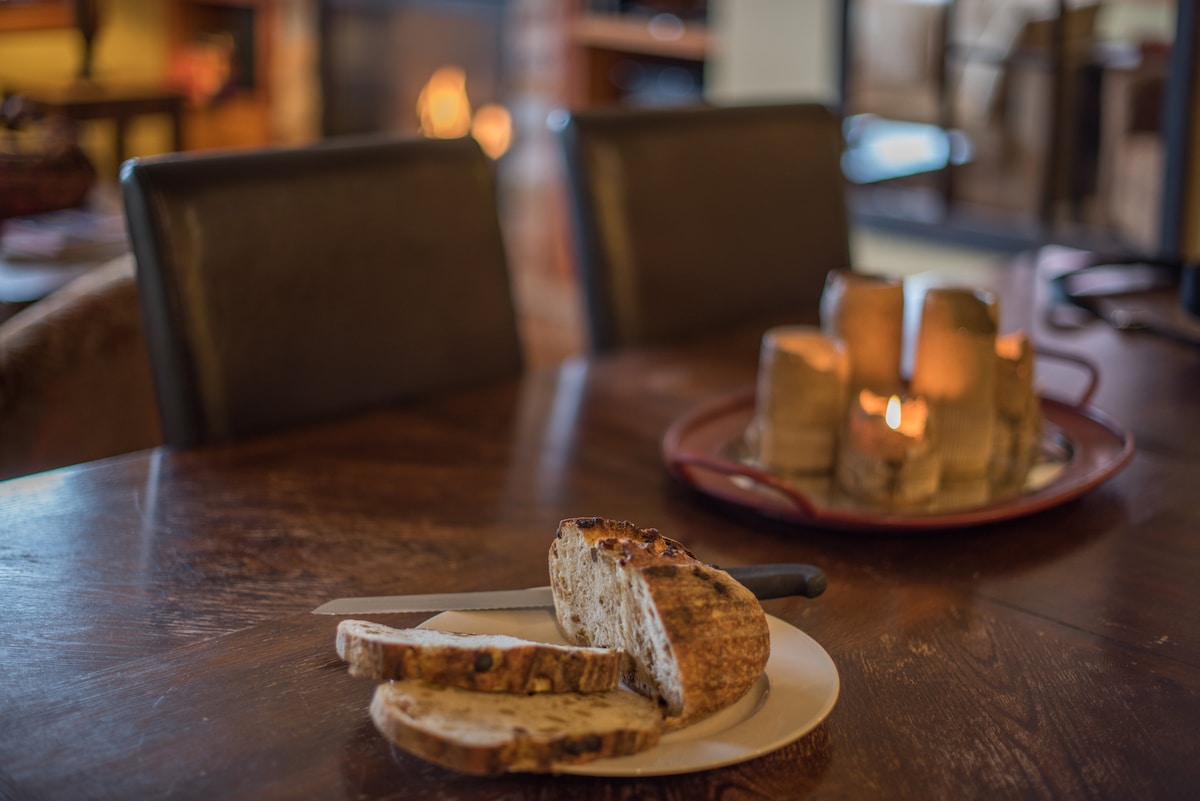 A wooden dining table is set with freshly sliced bread on a white plate. A knife rests beside the bread. In the background, a decorative platter with candles is softly lit, adding a warm ambiance to the dining space.