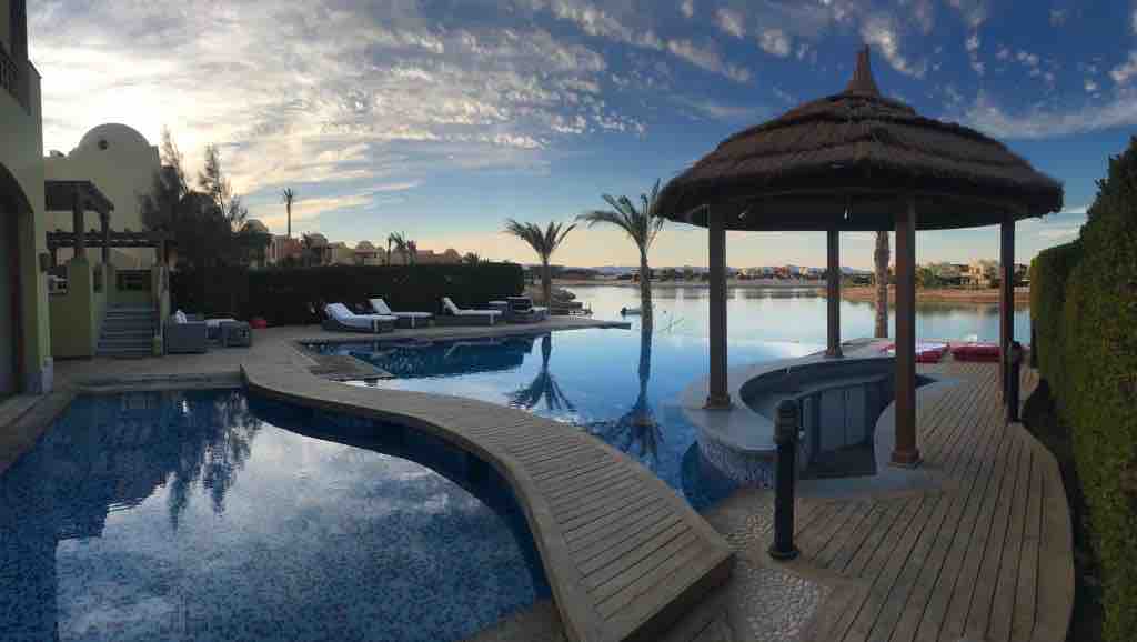 An outdoor pool area is surrounded by well-maintained greenery and palm trees. A thatched-roof gazebo offers seating near the water, with wooden decking leading to the pool. Sun loungers are arranged along the water's edge, reflecting a serene atmosphere with a clear sky above.