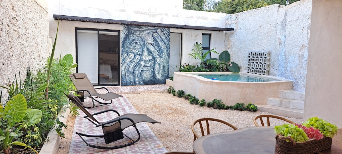 An outdoor space features a small pool surrounded by lush greenery. Two lounge chairs are positioned nearby, and a dining table with decorative plants is visible. A large mural of hands is displayed on the back wall, creating a striking focal point.