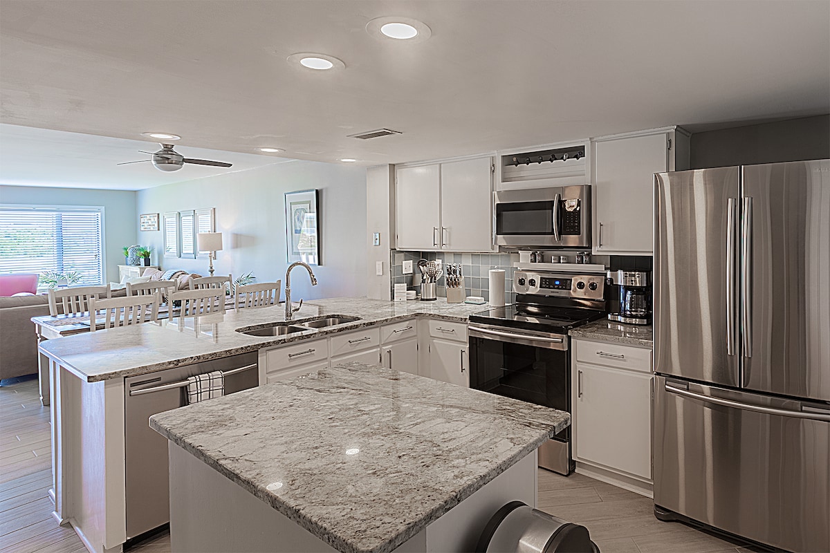 A modern kitchen features light cabinetry and granite countertops, complemented by stainless steel appliances. An island with seating invites casual dining. Natural light brightens the space, with the living area visible through an open layout. Ceiling lights provide additional illumination.