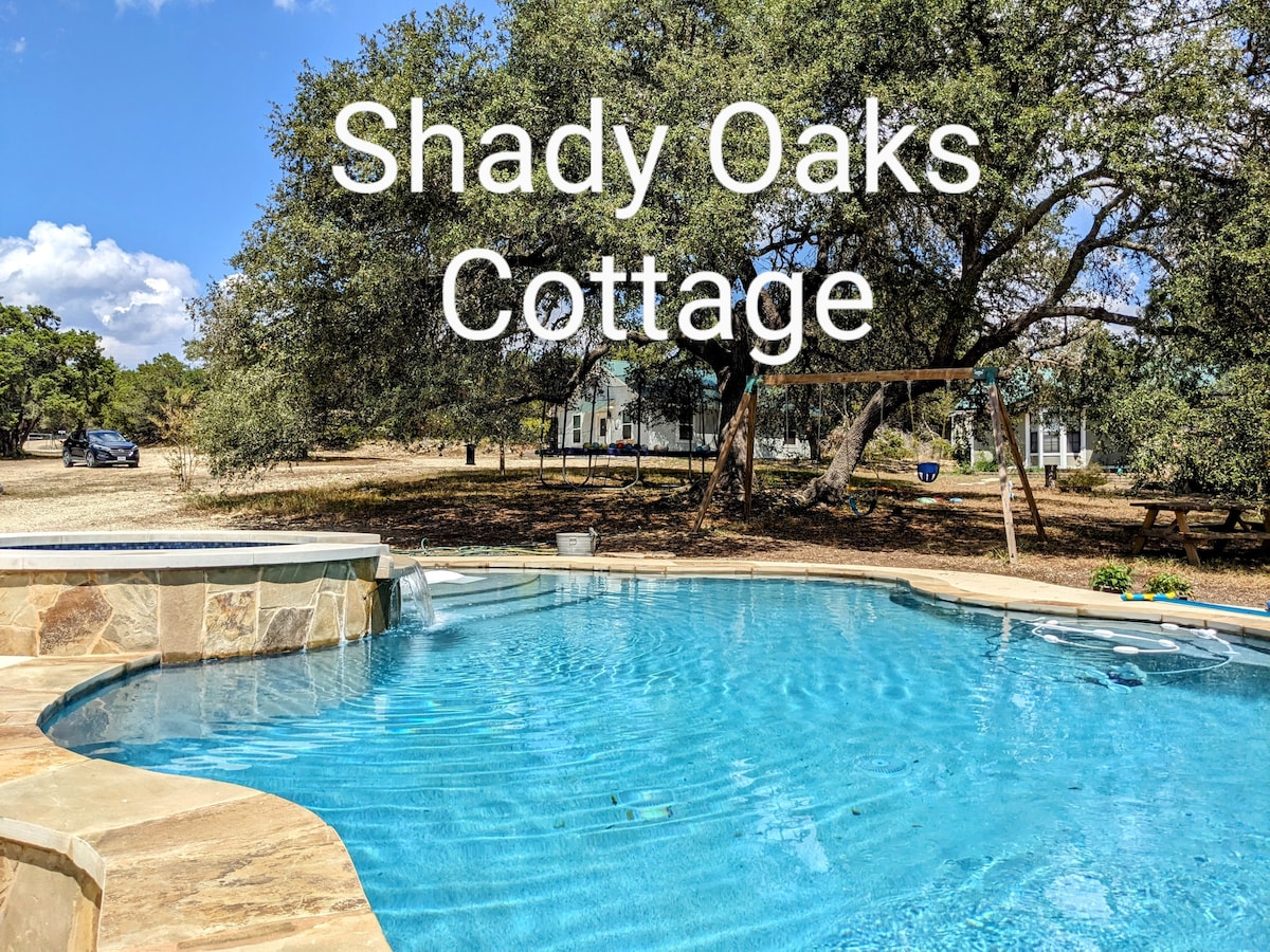 A large swimming pool is visible, surrounded by natural stone decking and shaded by oak trees. The water reflects the sky, enhancing the outdoor setting. In the background, a swing set and picnic tables can be seen, illustrating the spacious yard perfect for leisure activities.