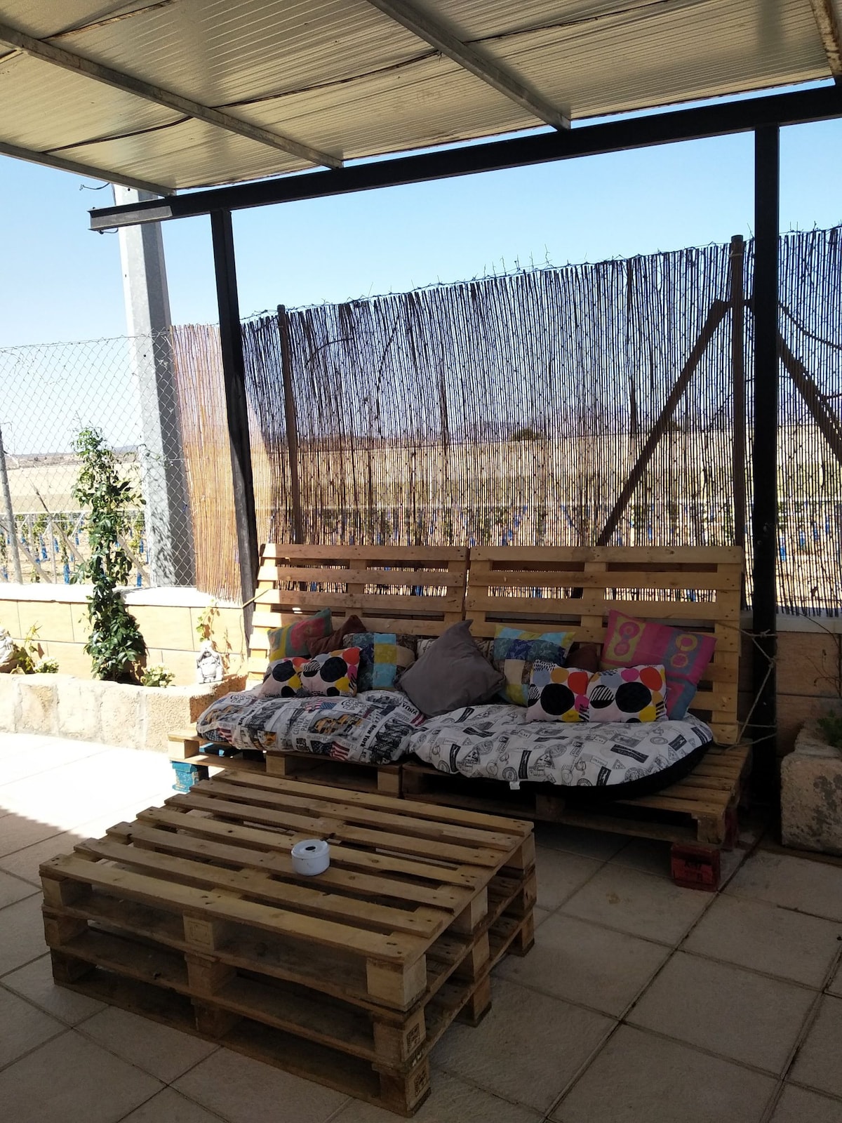 A designated outdoor seating area features a wooden pallet sofa adorned with colorful cushions and a patterned throw. A simple coffee table made from pallets is positioned in front. Natural light filters through a translucent roof, enhancing the space's relaxed vibe.