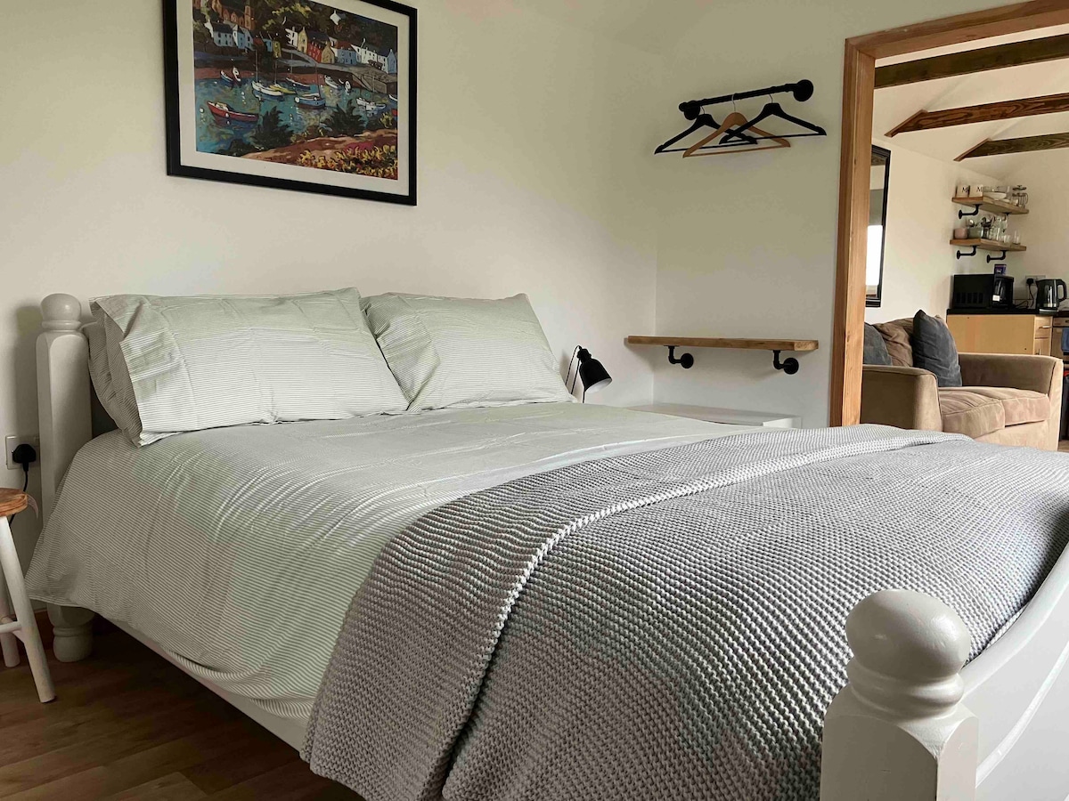 A spacious bed is dressed in soft linens, complemented by a grey knitted throw. A framed artwork hangs above the bed, and practical hooks are mounted on the wall. A small bedside table provides a resting place for essentials, with a cozy seating area visible in the background.