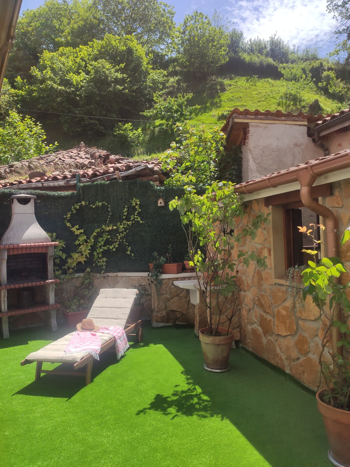 A private patio area is featured, showcasing a sun lounger on artificial grass. Stone walls provide a rustic backdrop, complemented by greenery and potted plants. A BBQ grill is visible, alongside a small washbasin, offering a cozy outdoor space for relaxation.