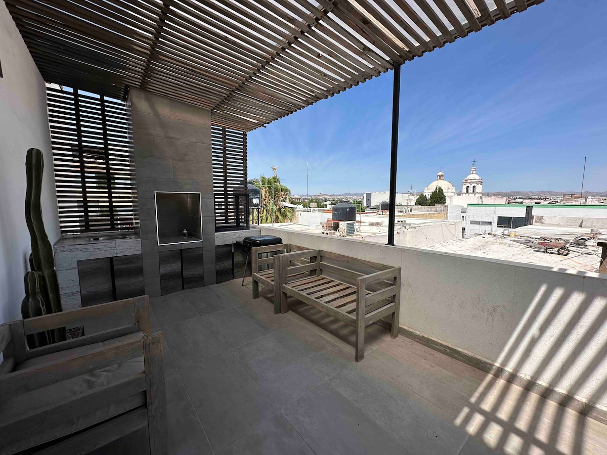 A spacious terrace offers a panoramic view of the surrounding area with wooden slats overhead providing shade. Minimalist seating arrangements are visible, complemented by a mounted TV on a textured wall. The distant skyline features architectural elements, creating an inviting atmosphere for relaxation.