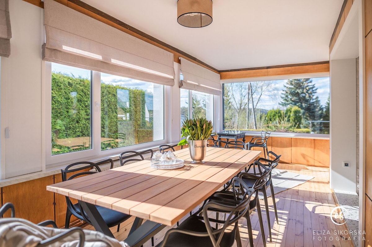 A bright dining area is featured, highlighted by large windows that offer views of the surrounding greenery. A long wooden table is surrounded by black chairs, creating a spacious and inviting atmosphere for meals.