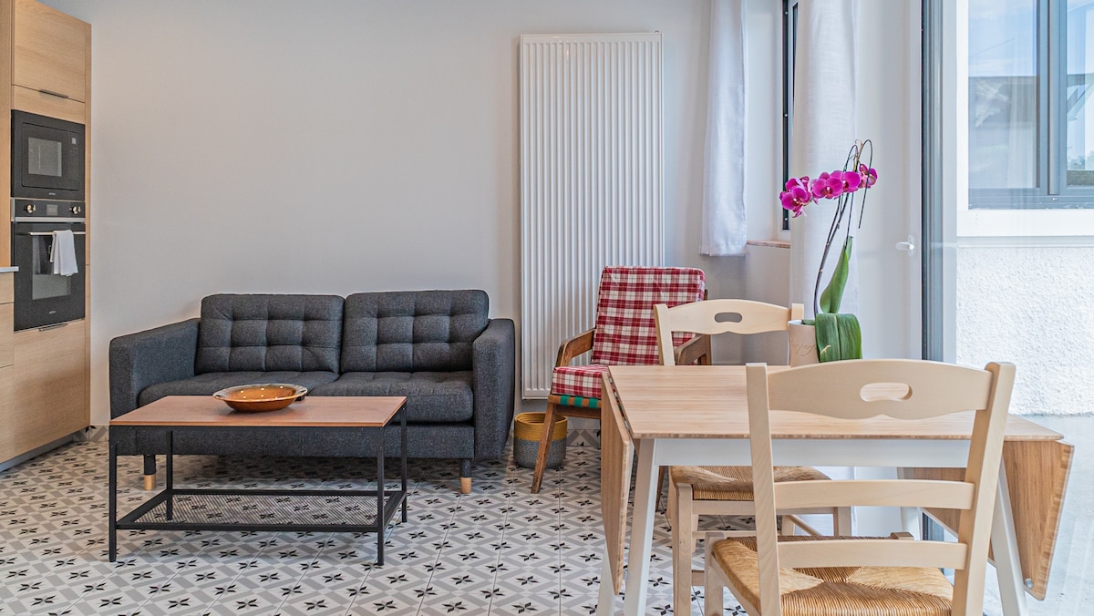 A modern living area is presented with a sleek gray couch and a stylish armchair featuring a checkered pattern. A simple wooden coffee table stands in front of the couch, complemented by a dining set with light-colored chairs. An arrangement of pink orchids adds a touch of color.