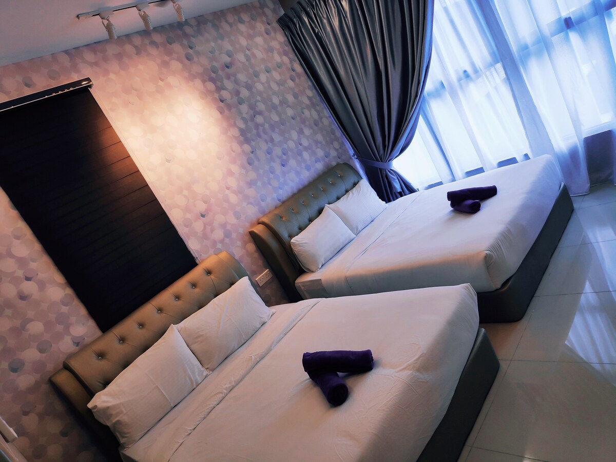 The bedroom features two spacious double beds adorned with fresh linens and towels. Soft purple accents add color to the decor, complemented by large windows allowing natural light to illuminate the room. Dark curtains frame the windows, providing an option for privacy.