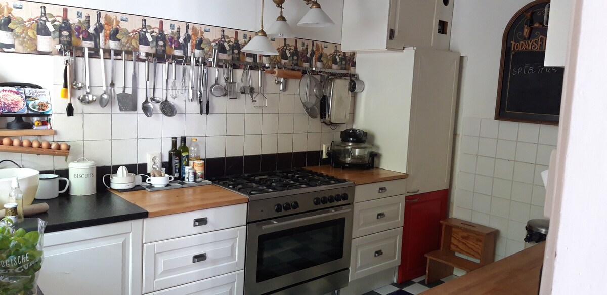 A well-equipped kitchen features a gas stove and an oven below a stainless steel hood. A countertop is lined with cooking utensils, oils, and a kettle. The walls are decorated with vintage-style tiles, and wooden shelves hold various jars and spices.