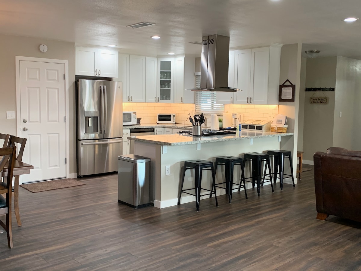 A modern kitchen is equipped with stainless steel appliances, including a refrigerator and oven. A large island with seating for four is complemented by sleek black bar stools. Soft lighting illuminates the granite countertops, and a living area is visible in the background.
