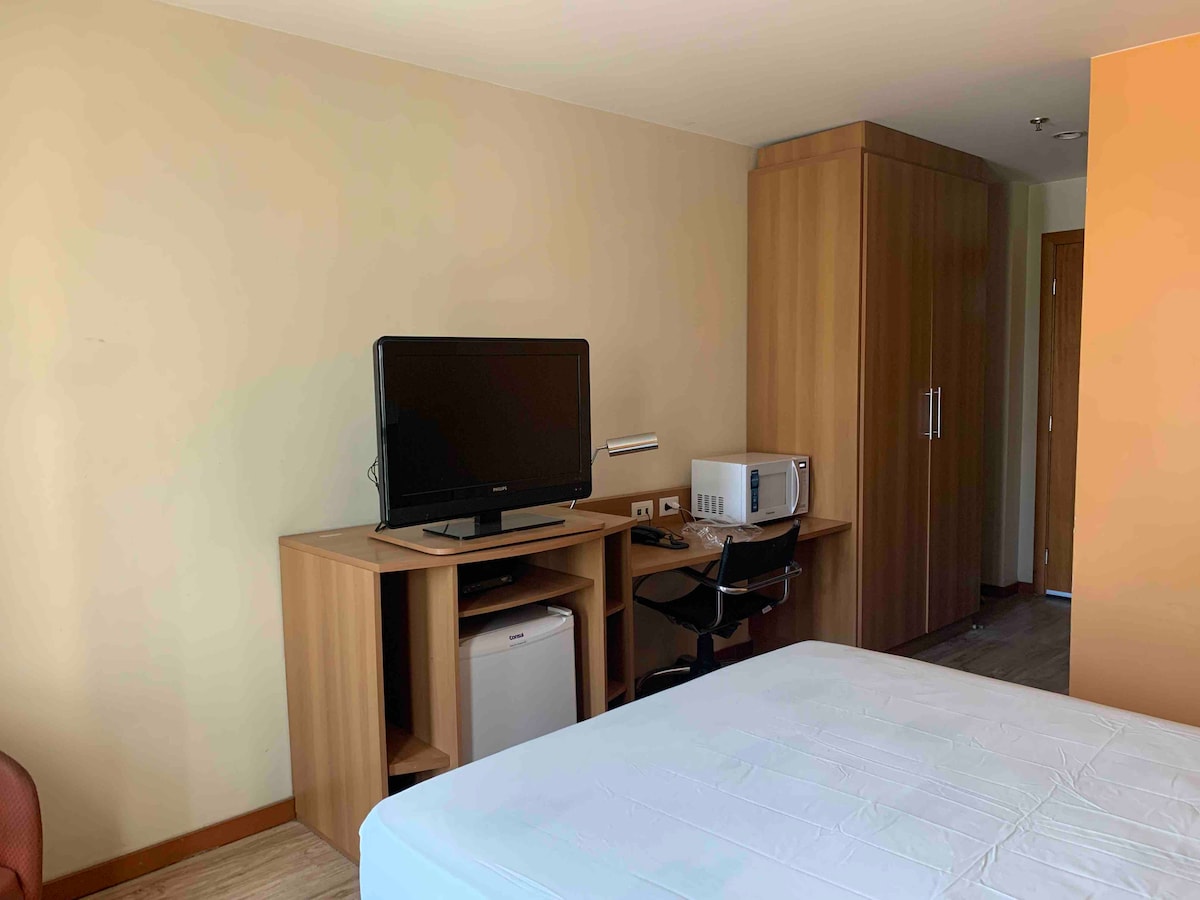 A spacious room features a queen size bed with a white mattress. A wooden entertainment unit includes a 32-inch TV and a microwave. An office chair is positioned at a desk, while a small refrigerator is beneath it. A wardrobe is visible on the right.