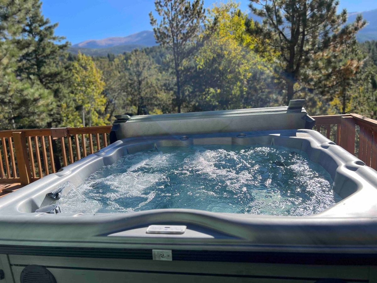 A hot tub is situated on a wooden deck, surrounded by towering evergreen trees. Views of the mountains are visible in the background, featuring a clear sky that enhances the peaceful setting. Water bubbles gently, inviting relaxation.