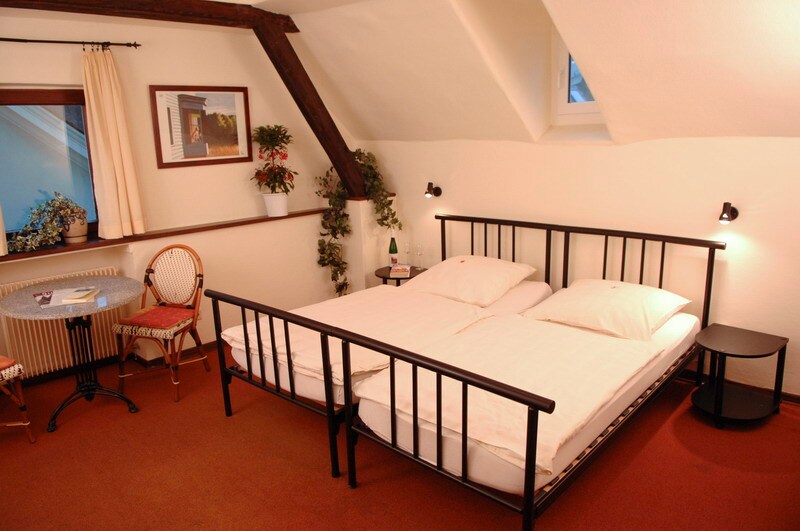 A cozy room features a spacious bed with white bedding, accompanied by a small round table and two chairs. Natural light enters through a small window, highlighting the wooden beams on the ceiling. A plant in the corner adds a touch of greenery to the space.