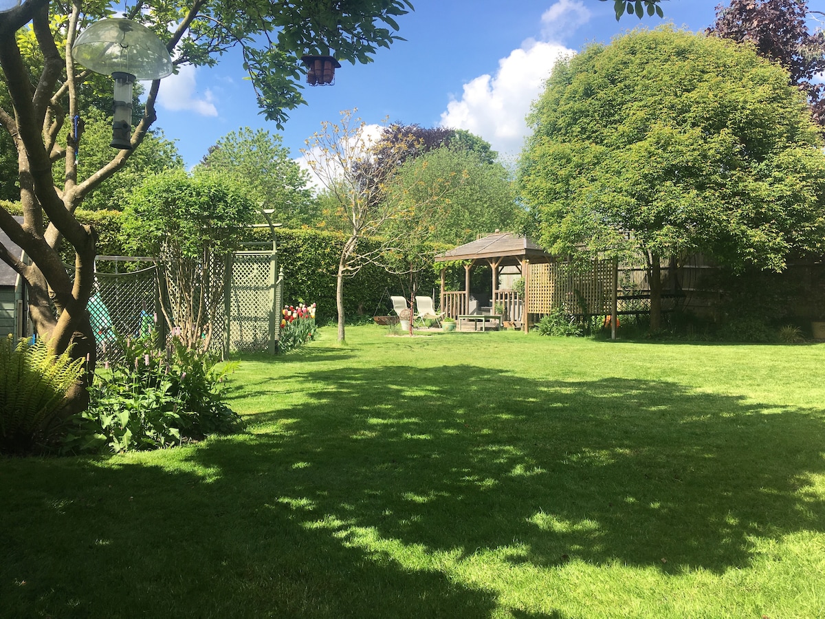 The spacious garden features a well-maintained lawn with diverse greenery and scattered trees. A wooden gazebo is visible in the background, accompanied by lounge chairs and colorful flower beds. Natural light filters through the branches, creating a peaceful outdoor ambiance.