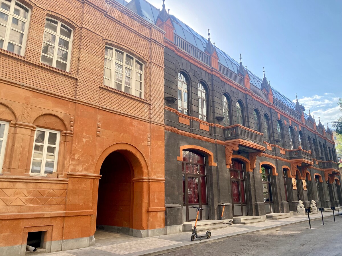 The image displays a newly renovated building with a blend of orange and dark stone facades, showcasing intricate architectural details. Large windows with white frames punctuate the exterior, and an arched entrance can be seen. The street is paved, and a scooter is parked nearby.